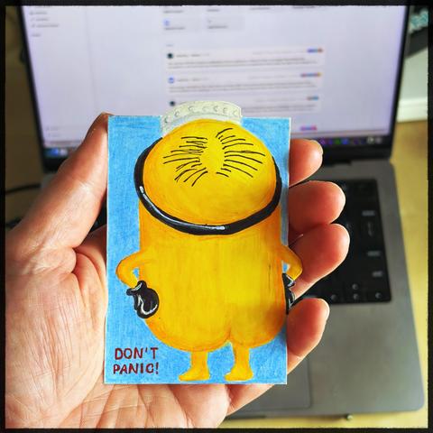 A person holding a card with a yellow cartoon (a Minion) character wearing a black belt and the words "Don't Panic!" in front of a laptop.