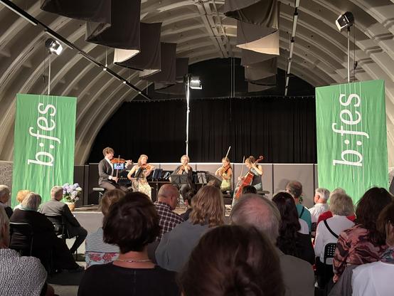 Ein Streichquartett spielt auf einer Bühne mit grünen Bannern, vor einem Publikum in einem gewölbten Raum.