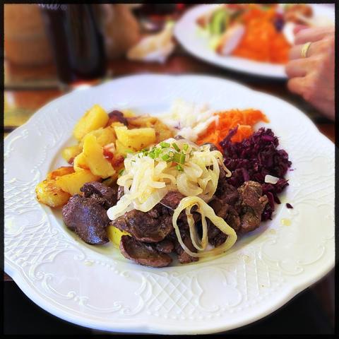 Ein weißer flacher Teller mit gebratener Hähnchenleber, glasierten Zwiebeln, Bratkartoffeln und Rohkostsalat. 