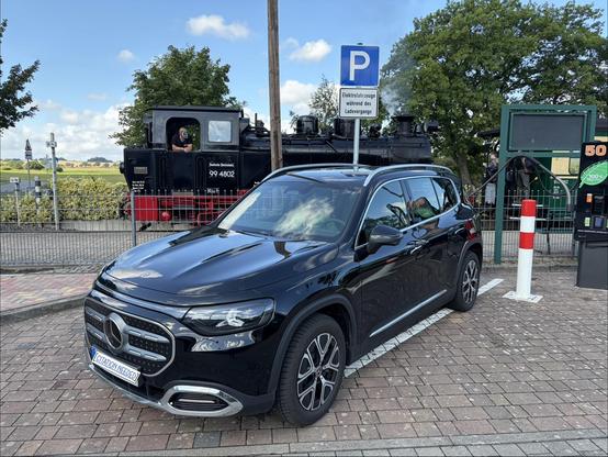 Ein Mercedes Elektro-SUV parkt an einer Ladesäule und lädt. Dahinter steht eine klassische Dampflok.