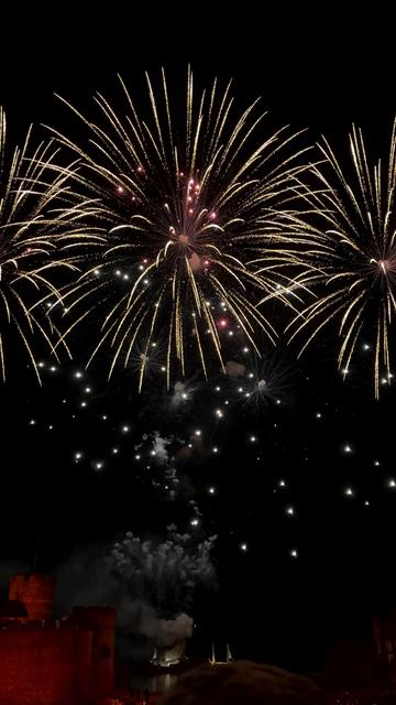 Das Bild zeigt einen nächtlichen Himmel, der von einem spektakulären, hellen Feuerwerk erleuchtet wird. Große goldene Effekte strahlen sternförmig nach oben, während darunter kleinere, funkelnde Lichtpunkte schweben. Unten links ist schemenhaft die rötlich beleuchtete Kulisse der bekannten Freilichtbühne zu erkennen.
