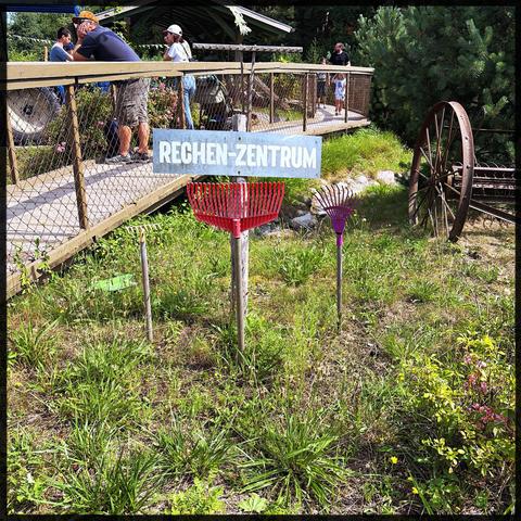 Ein Holzschild mit der Aufschrift "RECHEN-ZENTRUM" steht in einer grasbewachsenen Fläche, flankiert von bunten Rechenköpfen. Im Hintergrund sind Menschen auf einer Holzplattform in der Nähe einer Struktur zu sehen, mit viel Grün und einem Vintage-Rad.