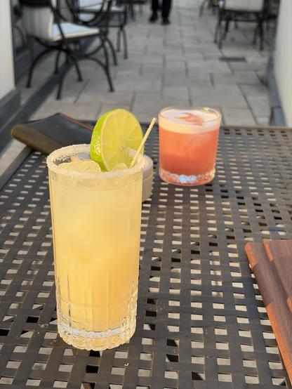 The image features two cocktails on a black wicker table. The foreground displays a tall, yellow drink garnished with a lime slice and a sugared rim. In the background, a shorter pink drink is visible, served in a clear glass. The setting