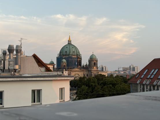 A view of a city skyline at sunset, featuring a prominent green domed building surrounded by trees, rooftops, and modern architecture in the background.