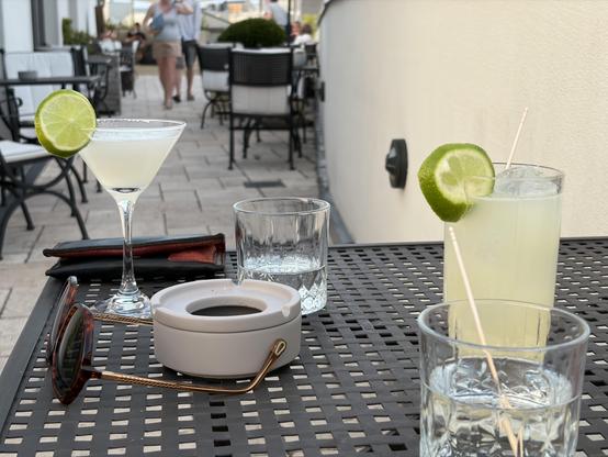 A table set for drinks with a view. Two cocktails with lime garnishes are featured: a martini and a tall glass. A few empty glasses and a pair of sunglasses are also on the table. The background shows a patio with people seated at