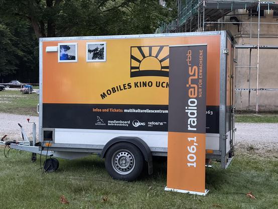A mobile cinema trailer with an orange and black design, featuring the words "Mobiles Kino UCK" and a logo with a sun. A standing banner displays the logo of "radioeins" along with the frequency "106.1." The trailer