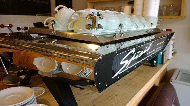 Eine "spirit" triplet Espresso-Maschine mit Chrome finish und einer schwarzen Frontplatte, sowie schwarzen Füßen. Oben auf der Maschine sind etliche Tassen gestapelt zum vorwärmen. Die Maschine ist auf einem Holztresen 