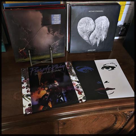 The "Purple Rain" Vinyl album sleeve sits on a wooden sideboard. The main picture shows prince on a motorcycle with his purple 80s suite. In the background some more vinyls of Raye "This music may contain hope" and Michael Kiwanuka "Love & Hate"