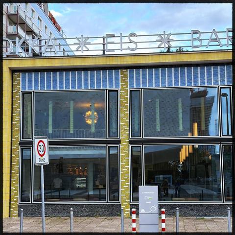 The image features a modern building with large windows, showcasing a stylish ice cream and bar establishment. The facade is decorated with yellow tiles and blue accents. A traffic sign indicating a speed limit of 30 km/h is visible in front, along with a