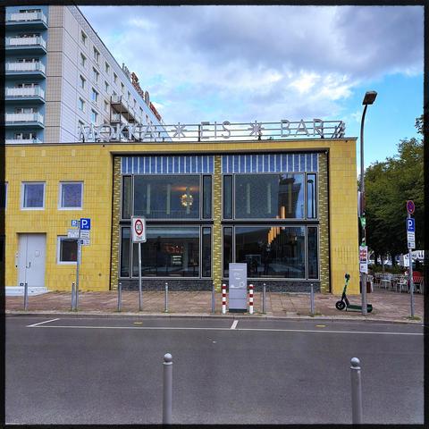 A modern ice cream bar named "Mokkai" is featured in the image. The building has a yellow tiled exterior with large windows. There are several parking signs visible, as well as a scooter parked outside. The scene is set against a cloudy