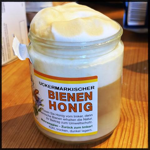 A jar of Uckermärkischer Bienenhonig (bee honey) sits on a wooden surface. The jar has frothy honey overflowing from the top and features a label with text in German promoting the natural benefits of honey and environmental conservation.