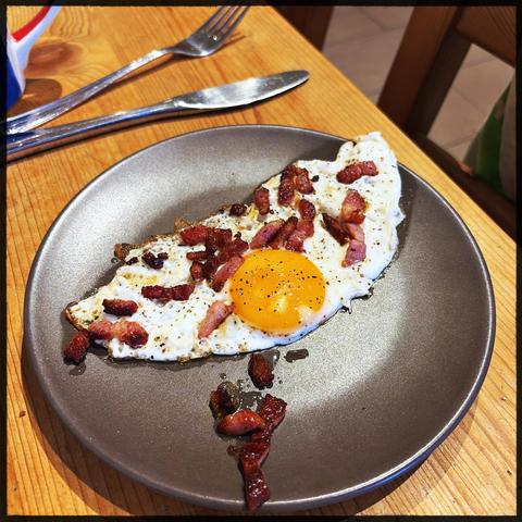 A fried egg with a runny yolk, topped with crispy diced bacon (pancetta-style) and cracked black pepper, served on a matte grey plate. Some extra bacon bits are scattered on the plate beside the egg. A fork and knife rest on a wooden table above the plate.