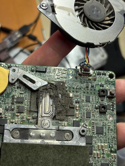 A hand is holding a computer motherboard with visible components, including a fan and a heat sink covered in dust. Various circuit elements and wires are seen on the board, indicating disassembly for cleaning or inspection. Mount under the fan is completely covered with greased dust. 