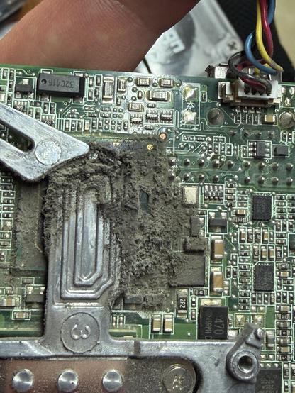 A close-up view of a circuit board covered in dust and debris. Components such as chips and capacitors are visible, along with a metallic part. A finger is partially visible in the image, indicating that the board is being held.
