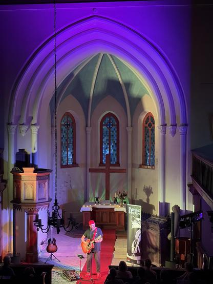 A musician performing on stage inside a church, illuminated by colorful lights. The background features stained glass windows, a wooden altar, and a cross. A guitar is placed on the floor, and an audience is seated, enjoying the performance.
