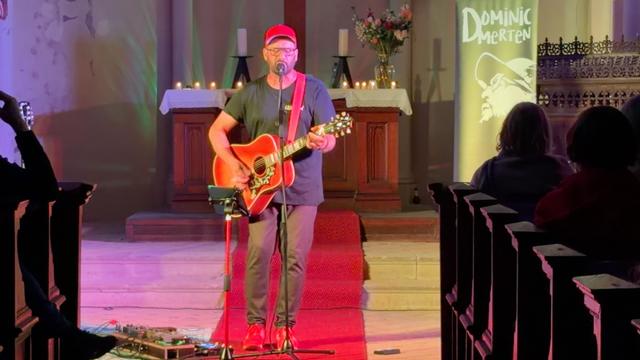 A musician performs on stage with an acoustic guitar in a church setting, wearing a red cap and black t-shirt. There are candles and a floral arrangement on the altar, and an audience is visible in the background. A banner displays the name "Domin