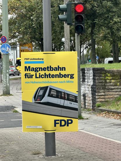Wahlplakat der FDP für Berlin 2026: Magnetbahn für Lichtenberg. Das Plakat zeigt eine Magnetschwebebahn auf einer Betonschiene auf gelbem Hintergrund 
