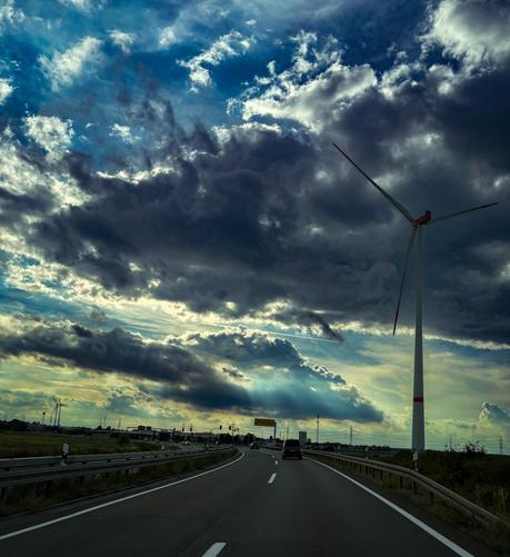 A scenic view of a highway curving towards the right, flanked by a wind turbine and moody clouds above. Sunlight breaks through the clouds, creating a dramatic sky, while vehicles are visible on the road.