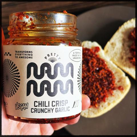 An open jar of "Pretty Noms Chili Crisp Crunchy Garlic," featuring a vibrant red topping, is held above a plate with two slices of bread coated in the chili crisp. The jar label emphasizes that the product is vegan and suggests it enhances flavors