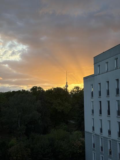 The setting evening sun radiating orange from behind the corner of an apartment building. Sun rays are visible in the clouds. In the left part of the picture a forest can be seen and behind that the upper part of the Berlin television tower 