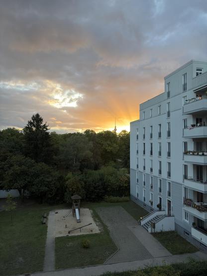The setting evening sun radiating orange from behind the corner of an apartment building. Sun rays are visible in the clouds. In the left part of the picture a forest can be seen and behind that the upper part of the Berlin television tower. In the bottom part a small playground with a slide and some lawn. 