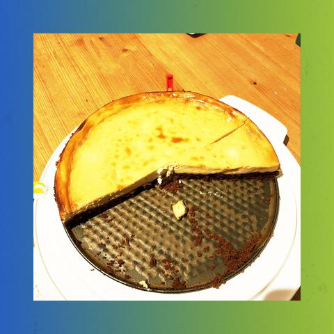 Ein New York Cheesecake auf einem weißen runden Kuchenteller. Der Kuchen ist schon angeschnitten und es fehlt schon fast die Hälfte. Die Oberfläche ist golden-braun. Der Kuchen befinden sich auf dem Boden einer Springform von welcher der Rand schon abgenommen wurde. Das ganze steht auf einem Holztisch 