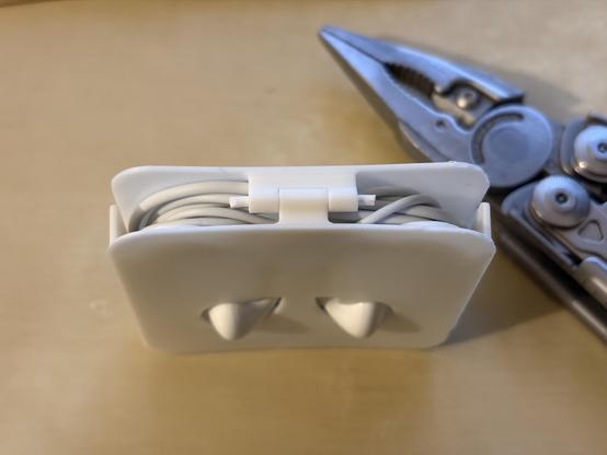 A 3D printed white plastic case for wired earpods, seen from above. The side is open, so the wound cables can be seen below a hinge. The hinge is fitted with a piece of filament. On the right side, a piece of Leatherman multi-tool is visible. 