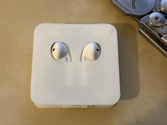 A white 3D-printed square plastic case for Apple wired earpods lying on a desk. The two pieces of the earpods are visible through cutouts. A partially visible Leatherman multi-tool in the upper right corner 