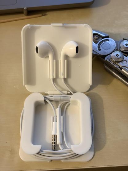 A white plastic case, 3D printed, for Apple wired earpods. The earpods are neatly folded inside the white case. In the background, partially visible, a leatherman multitool